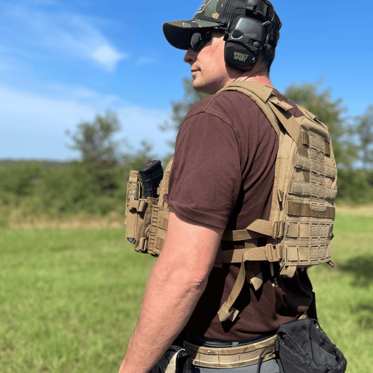Rear side of man wearing tactical gear including a vest, and ear protection in an outdoor setting.