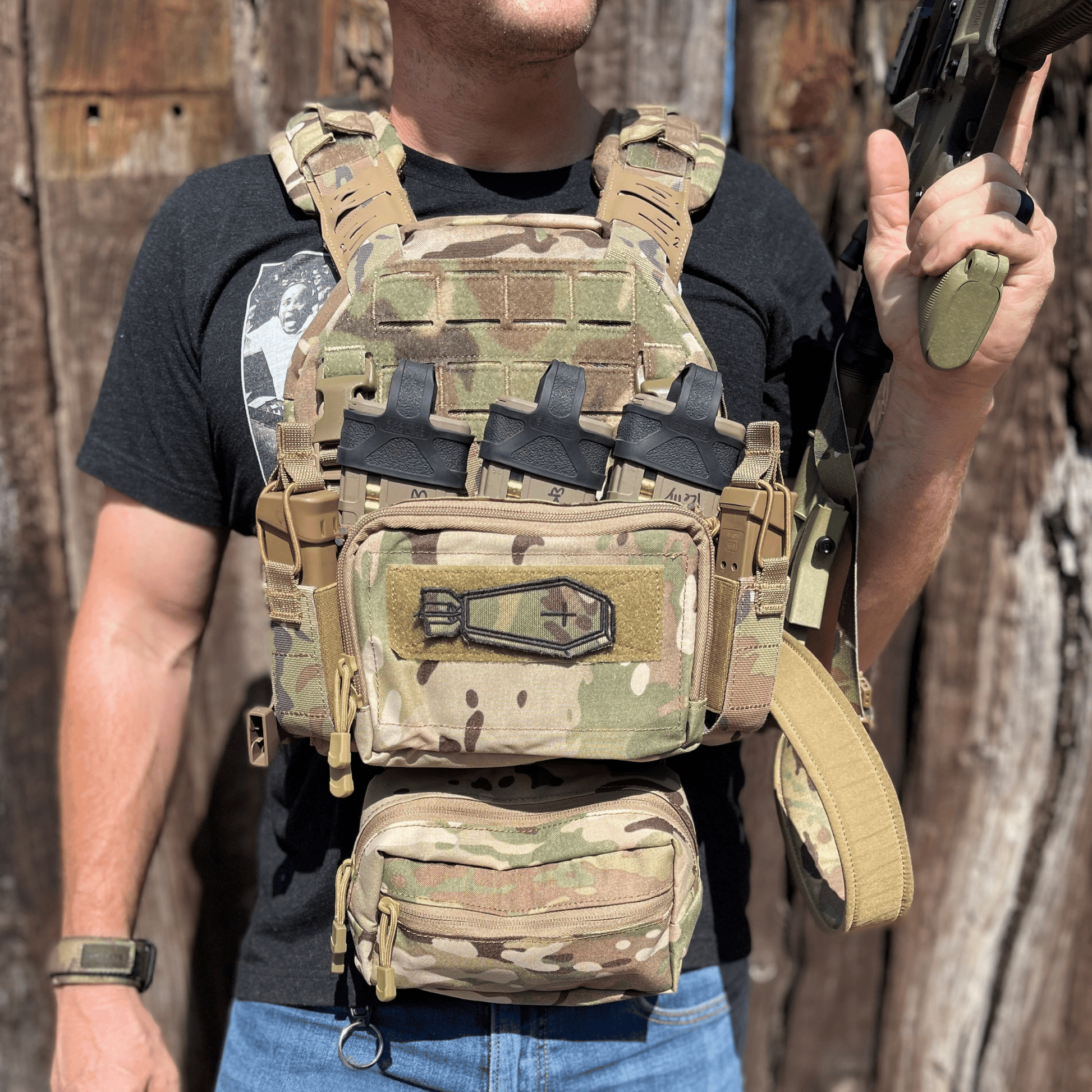Person wearing a camouflage tactical vest holding a rifle against a wooden background