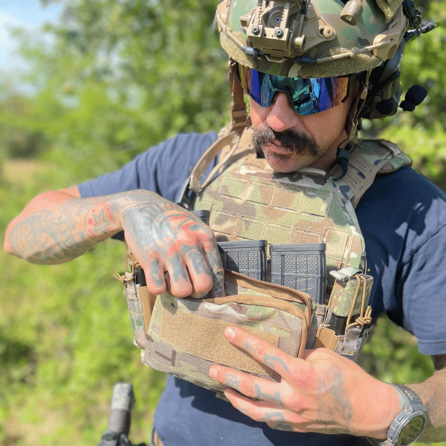 Person in tactical gear with a camouflage vest and helmet outdoors reaching into chest rig pouch.