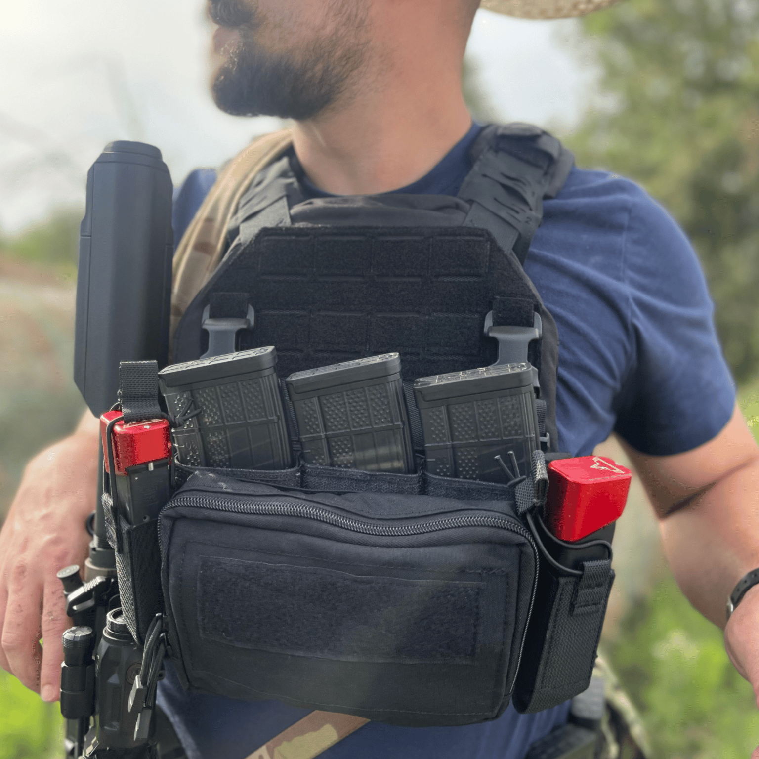 Person wearing a black tactical vest with multiple pouches and equipment outdoors